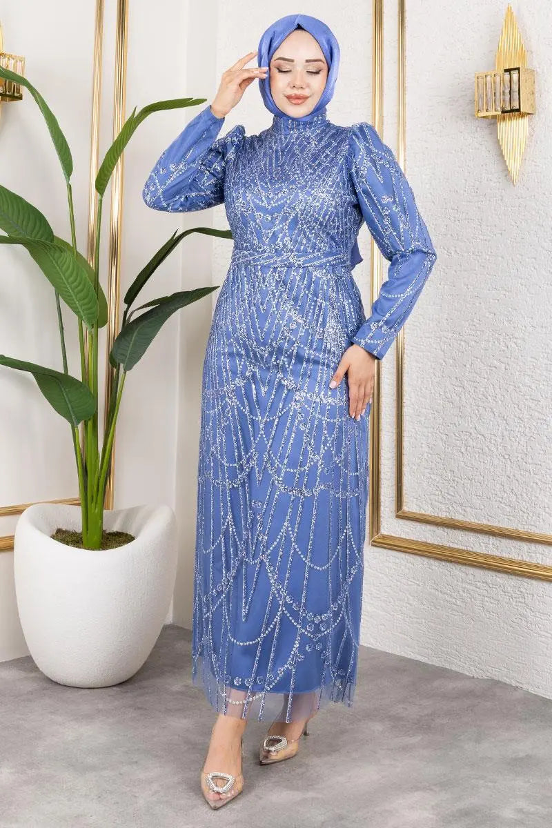 Woman in a blue dress with a hijab standing in a room with a plant and framed picture.