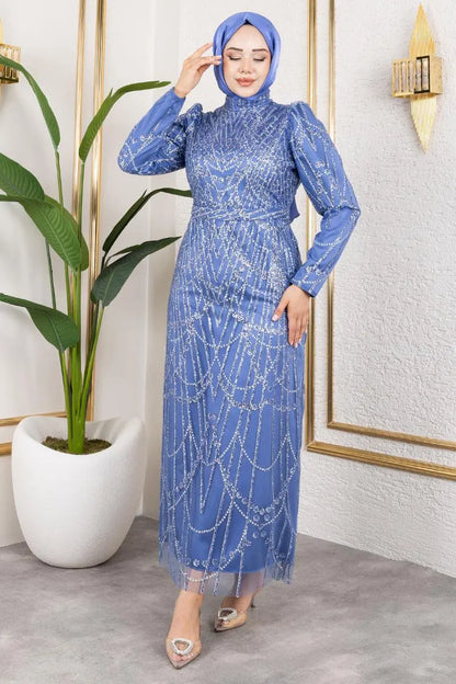 Woman in a blue dress with a hijab standing in a room with a plant and framed picture.