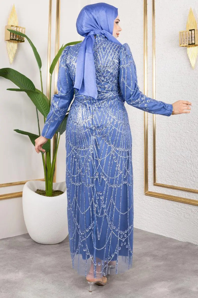 Woman in a blue dress with intricate patterns standing in a room with a plant and decorative wall.