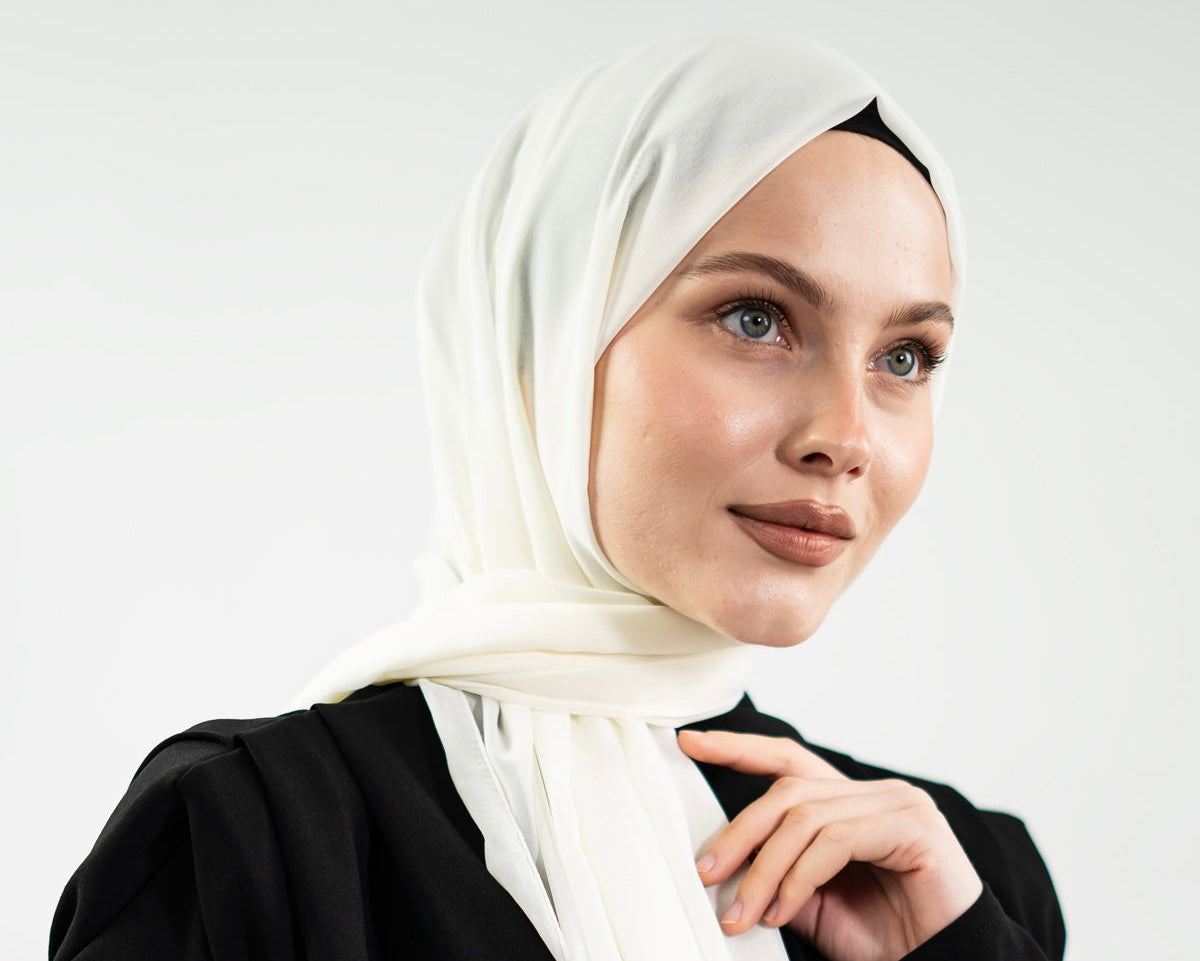 A woman wearing a white hijab and a black abaya, photographed against a clean light gray studio background.